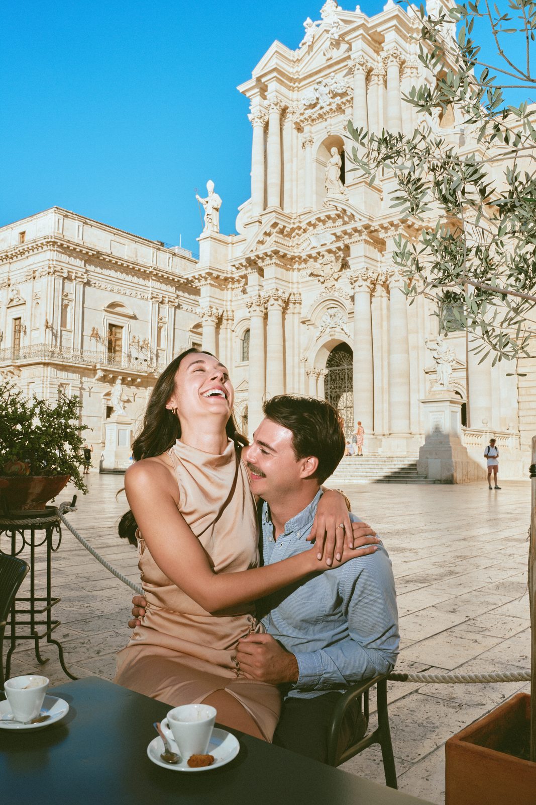 Couple laughing over coffee at a cafe in Piazza Duomo, the cathedral of Syracuse behind