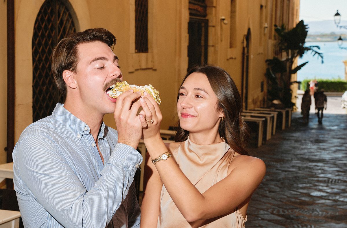 He takes a bite of the cannolo she is holding, in a street of Ortigia