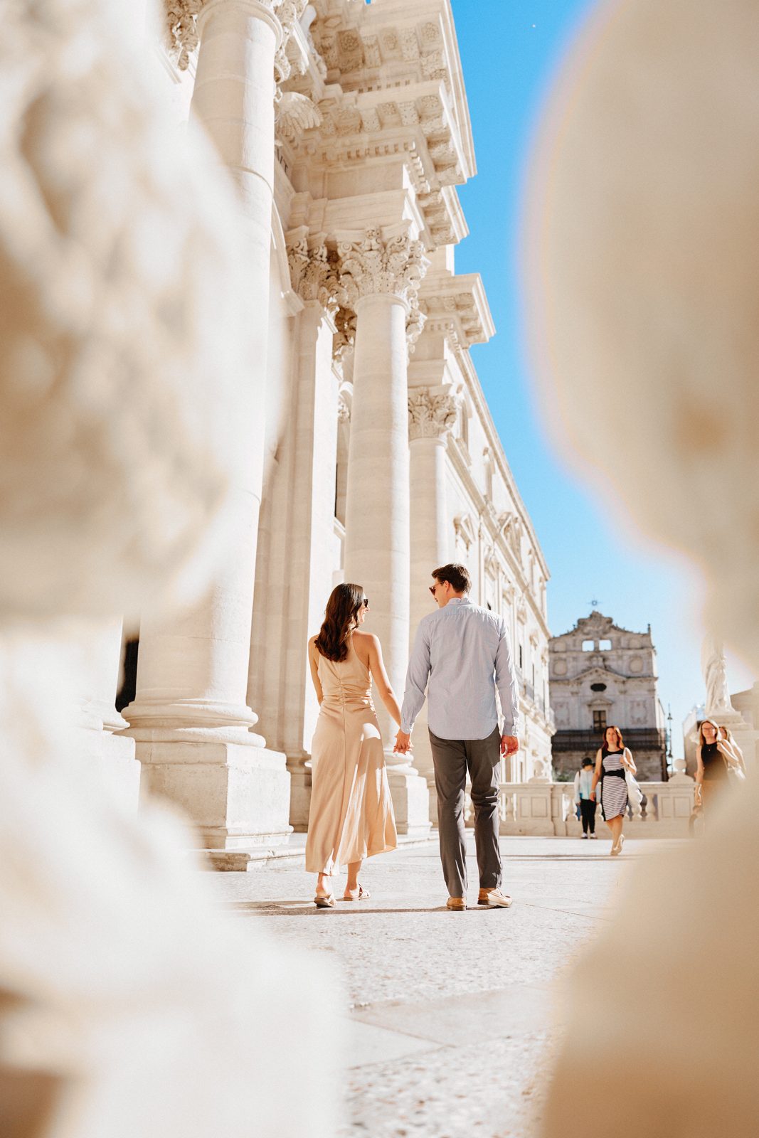 Couple walking hand in hand along the columns of the cathedral of Syracuse, seen through a stone balustrade