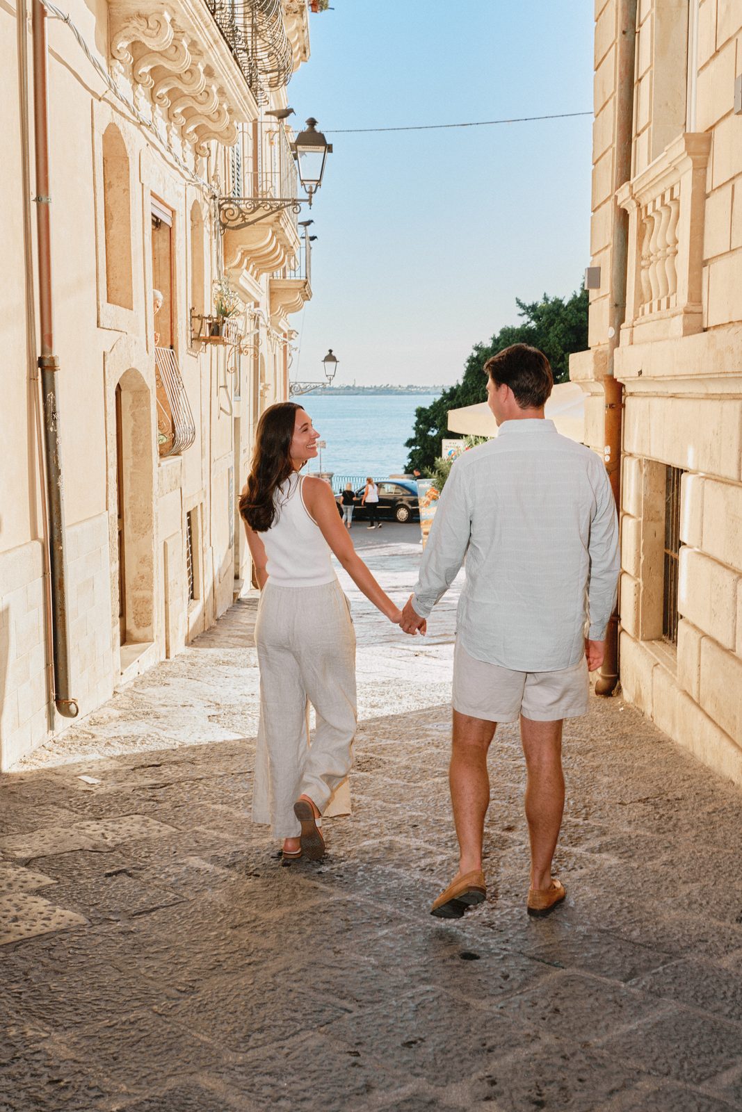 Couple walking hand in hand down a street of Ortigia towards the sea